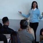 A confident woman gives a speech in front of a diverse audience at a Speakers Express Böblingen meeting, standing next to a whiteboard and engaging participants in an interactive Toastmasters session.
